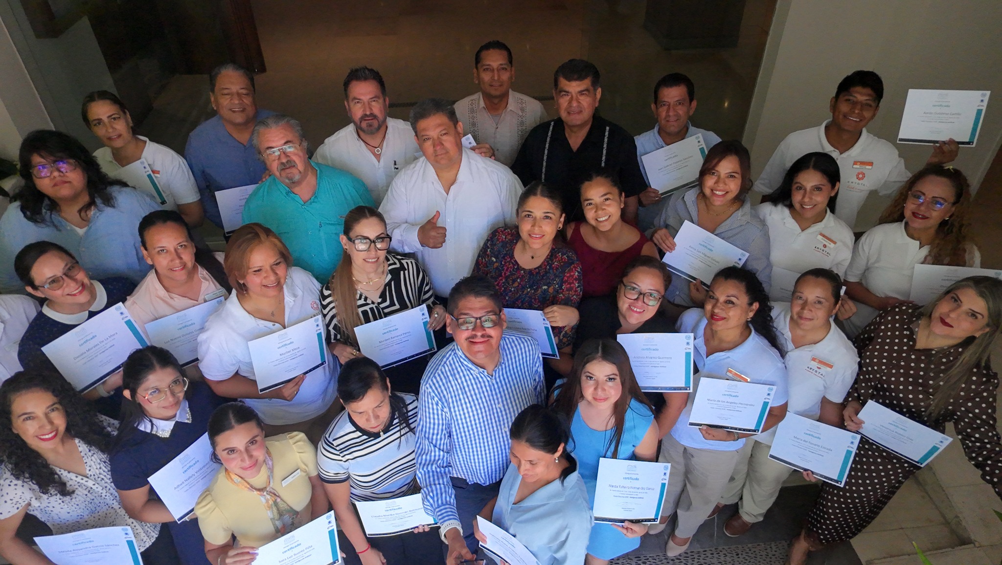 Participantes de capacitación hotelera reunidos en grupo, mirando hacia arriba y mostrando sus constancias